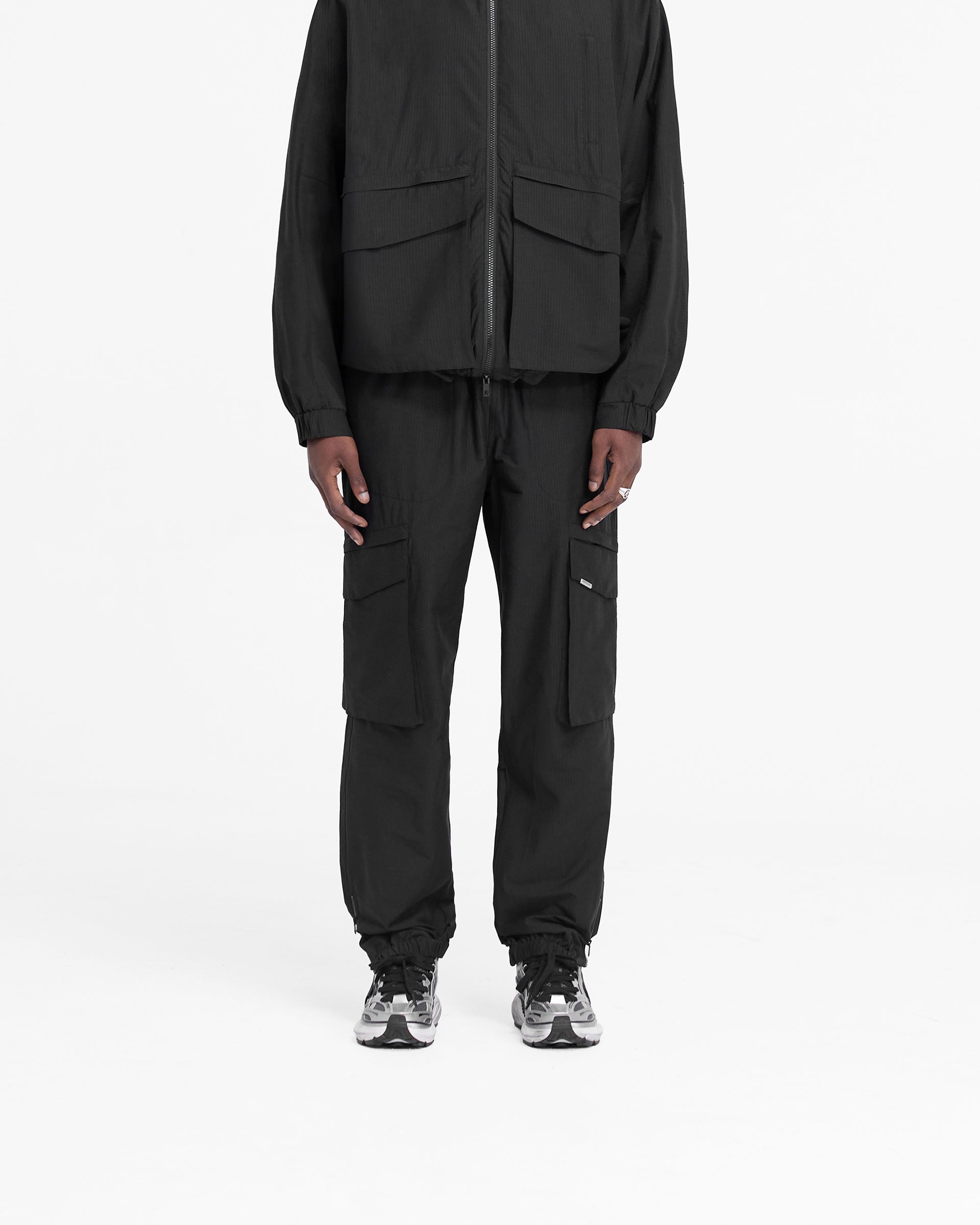 Man wearing black cargo pants on a plain white background | Tech Cargo Pant in Jet Black | Relaxed fit cargo pants with elasticated cuff and zip closure | FW24 Pants | Represent Clo