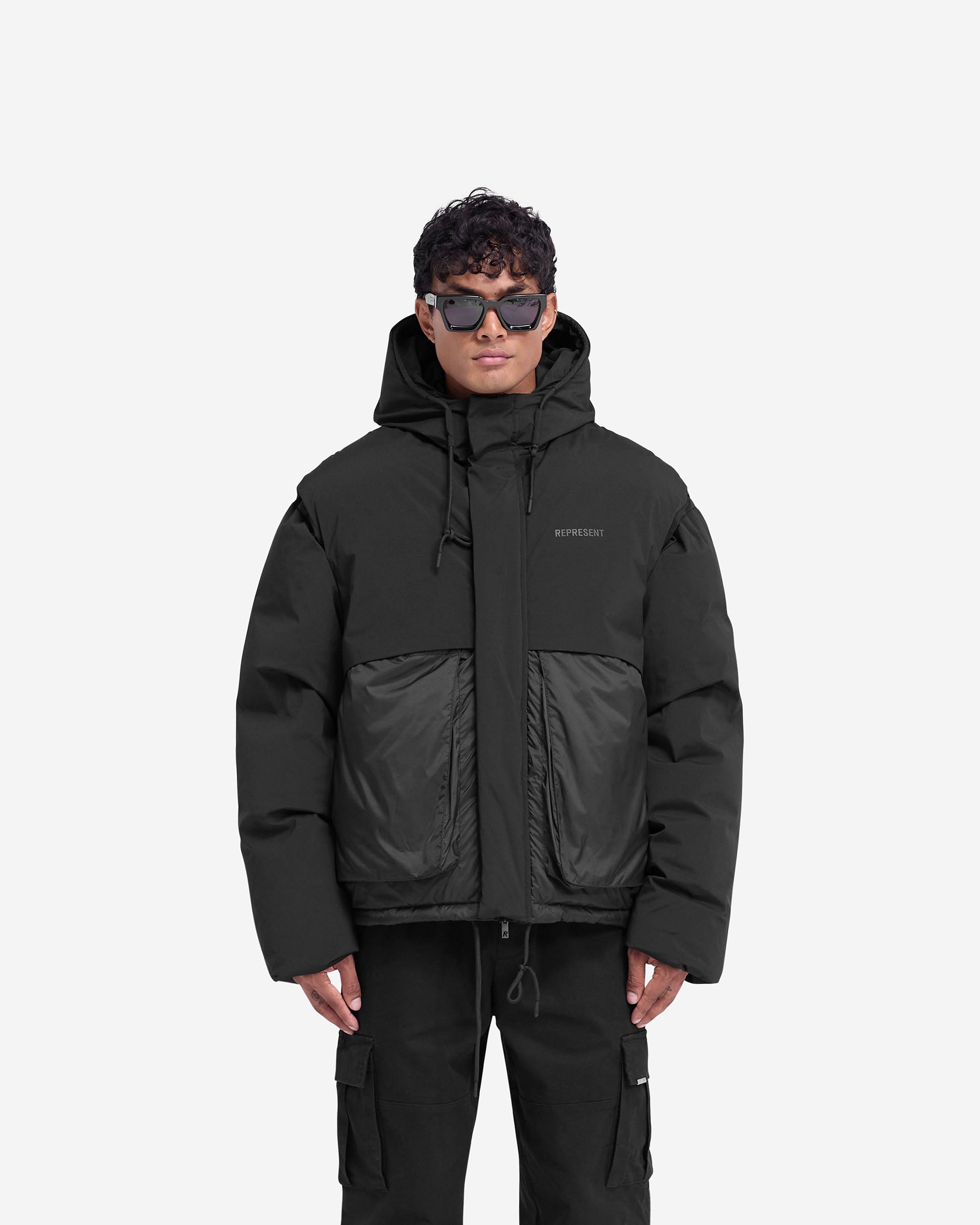 Man wearing a black puffer jacket with a hood on a plain white background | Layered Hooded Puffer in Jet Black | Duck down filled jacket with adjustable hood | FW24 Outerwear | Represent Clo