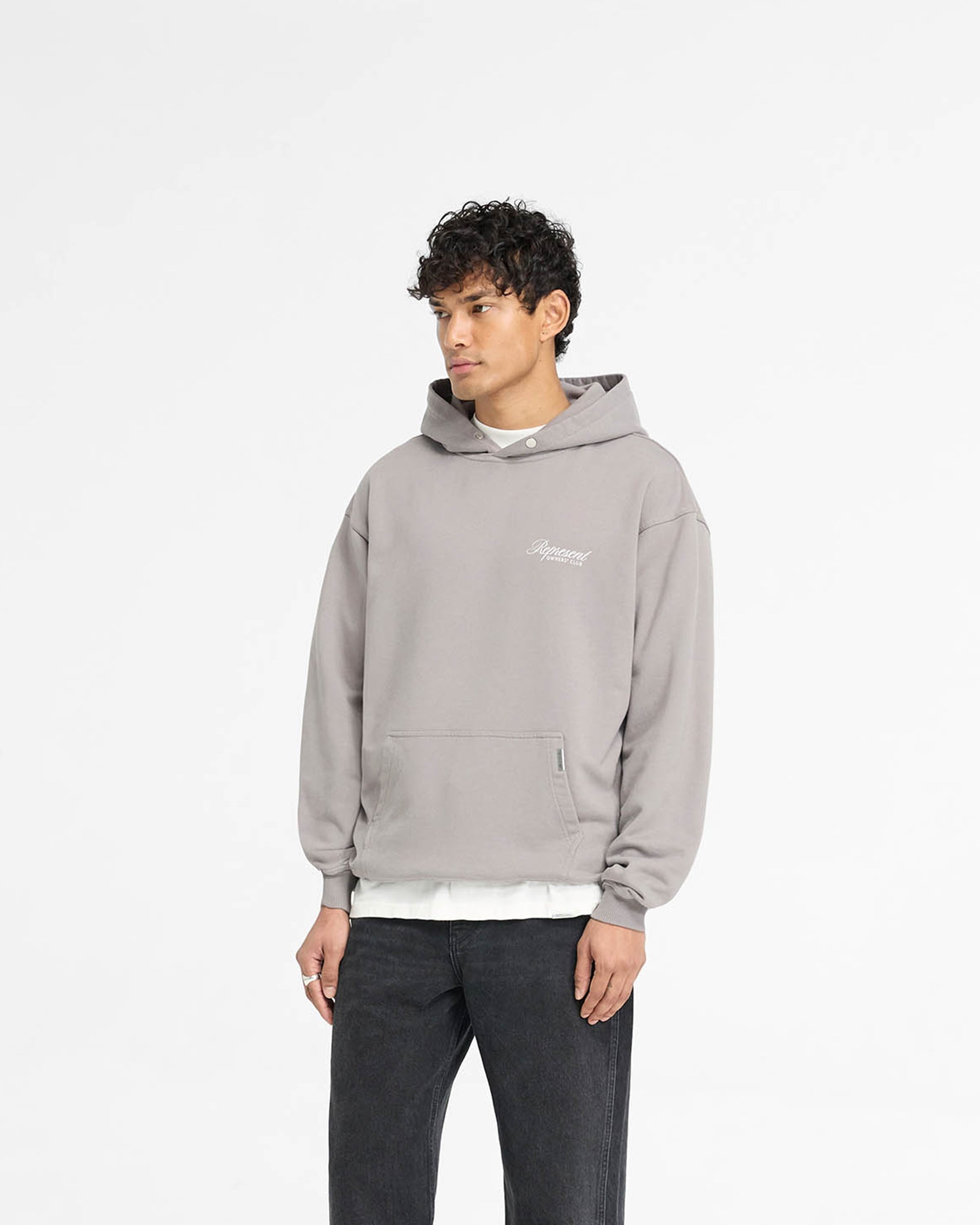 Man wearing a grey oversized hoodie on a plain white background | Represent Owners Club Script Hoodie in Slate | Heavyweight cotton hoodie with script graphic and Cobrax popper | Owners Club Hoodies | Represent Clo