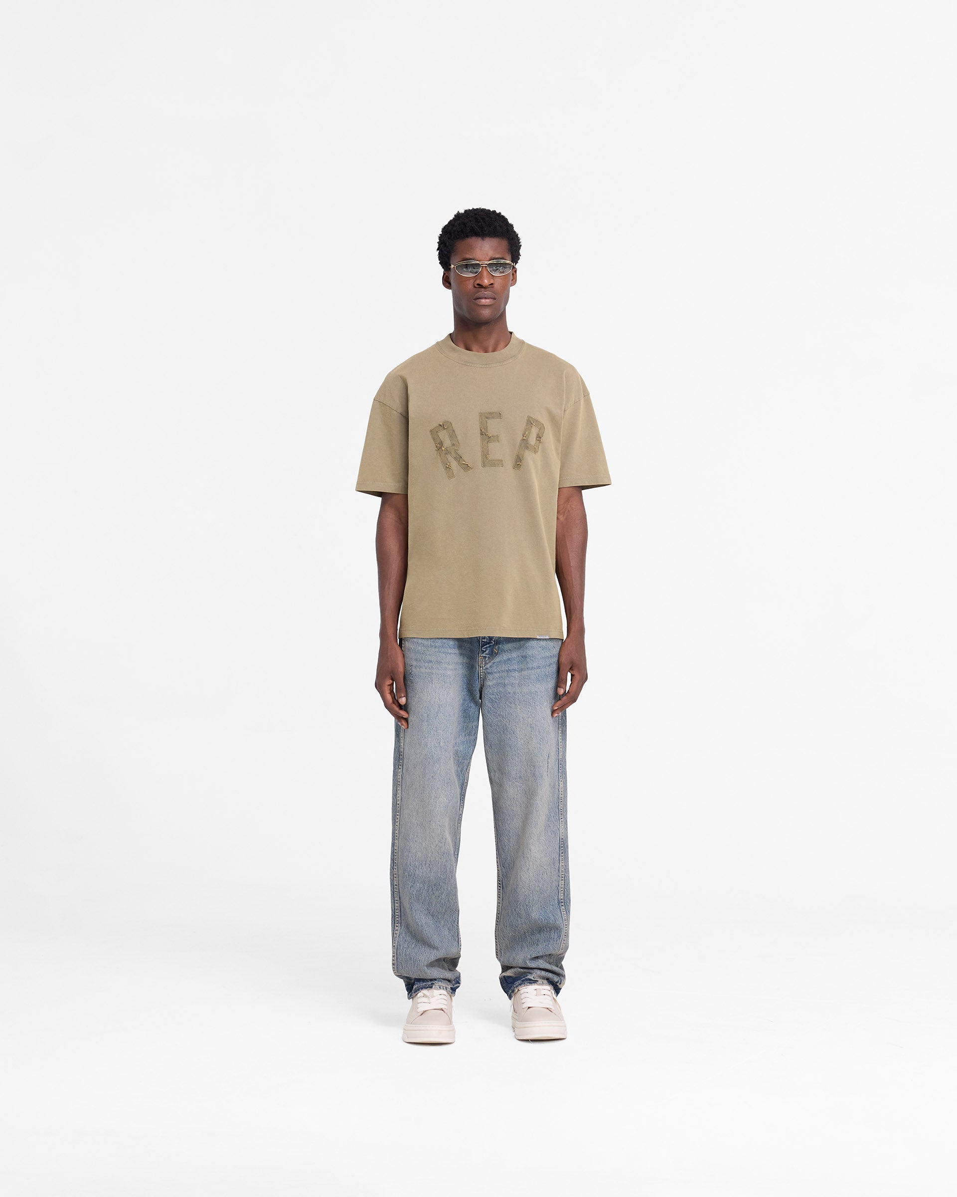 Man wearing a fawn t-shirt on a plain white background | Rep Applique T-Shirt in Fawn | Oversized t-shirt with distressed applique logo | FW24 T-Shirts | Represent Clo