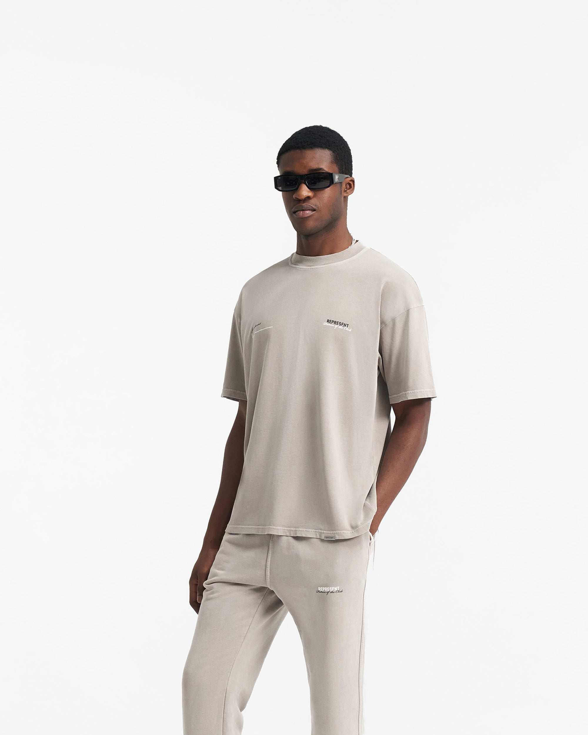 Man wearing a brown t-shirt on a plain white background | Patron Of The Club T-Shirt in Mudstone | Oversized t-shirt with club branding and ribbed crew neck | Patrons T-Shirts | Represent Clo
