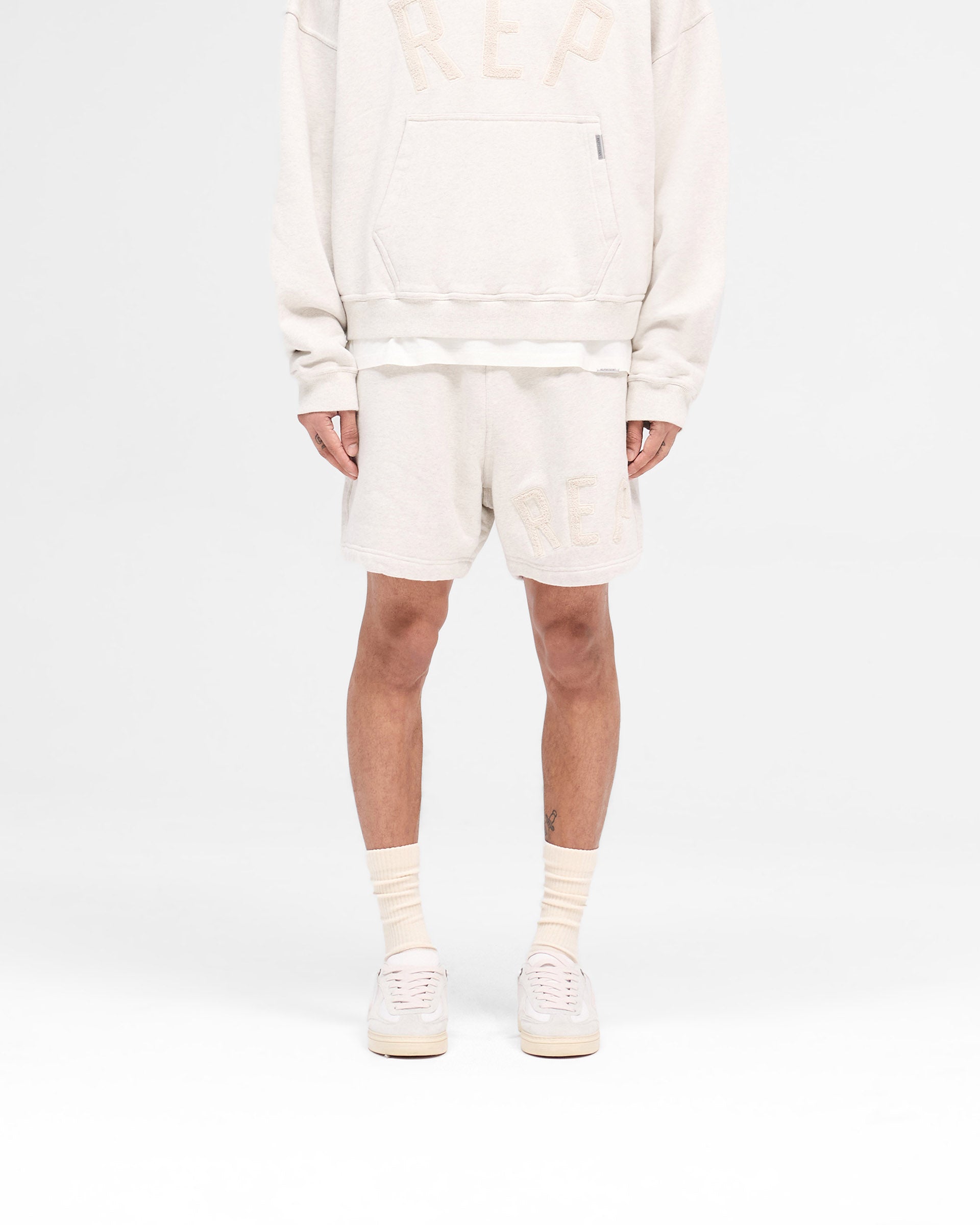 Man wearing cream shorts on a plain white background | Rep Applique Shorts in Cream Marl | Boxy fit shorts with elasticated waistband and applique logo | SS25 Shorts | Represent Clo