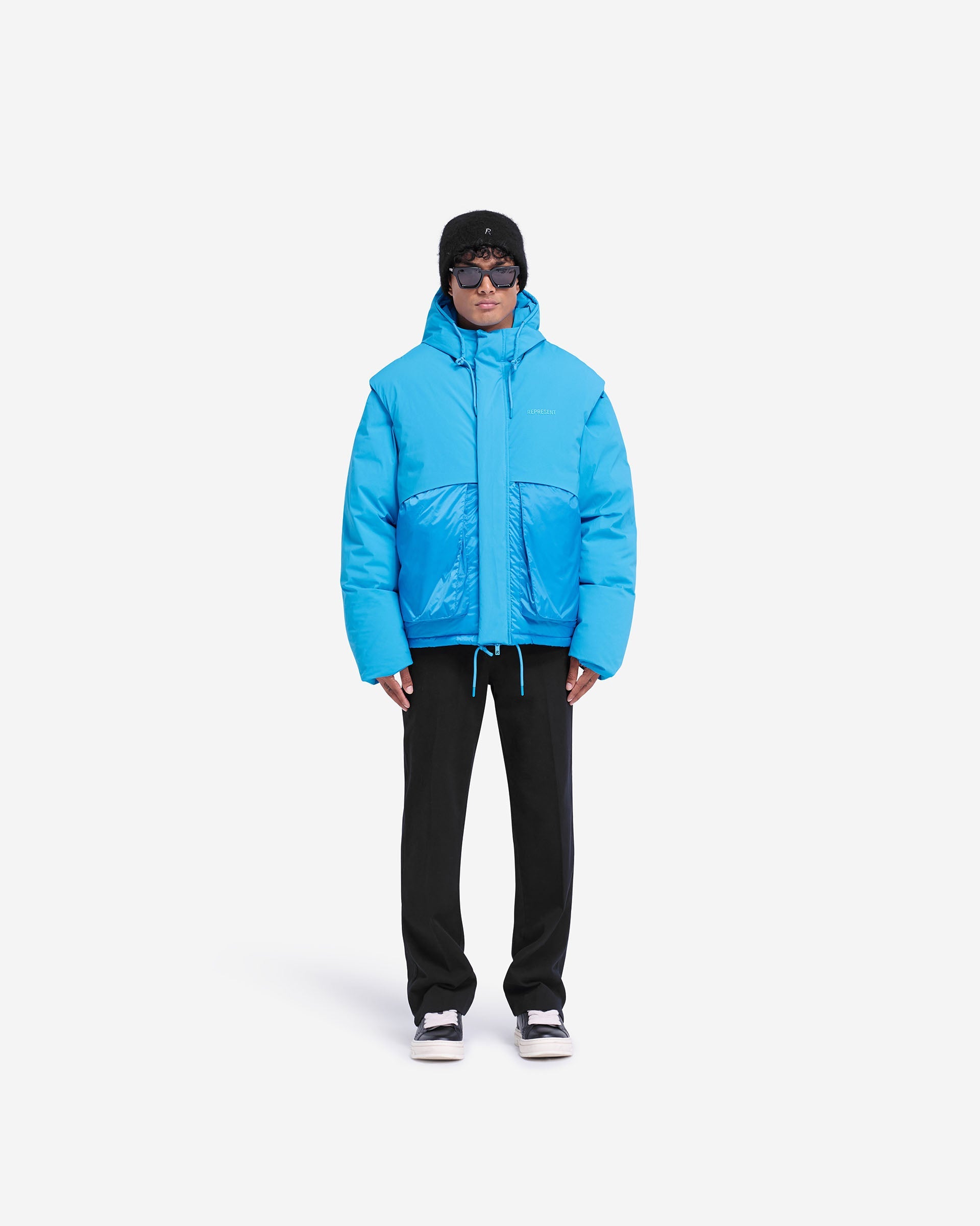 Man wearing an electric blue puffer jacket on a plain white background | Layered Hooded Puffer in Electric Blue | Duck down jacket with adjustable hood | FW24 Outerwear | Represent Clo
