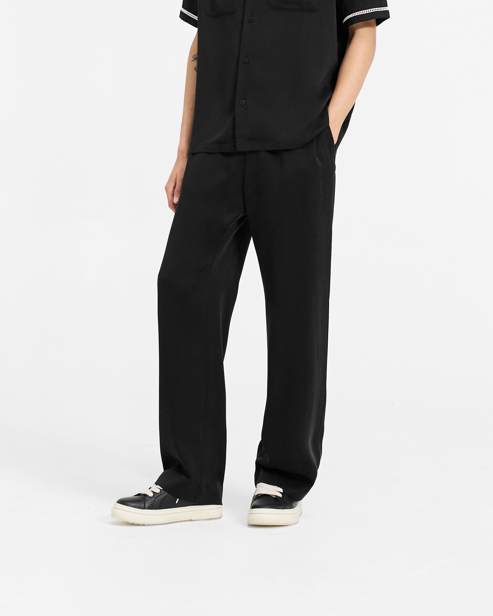 Man wearing black straight leg pants on a plain white background | Resort Pant in Jet Black | Drapey pants with elasticated waistband and ladder trim | SC24 Pants | Represent Clo