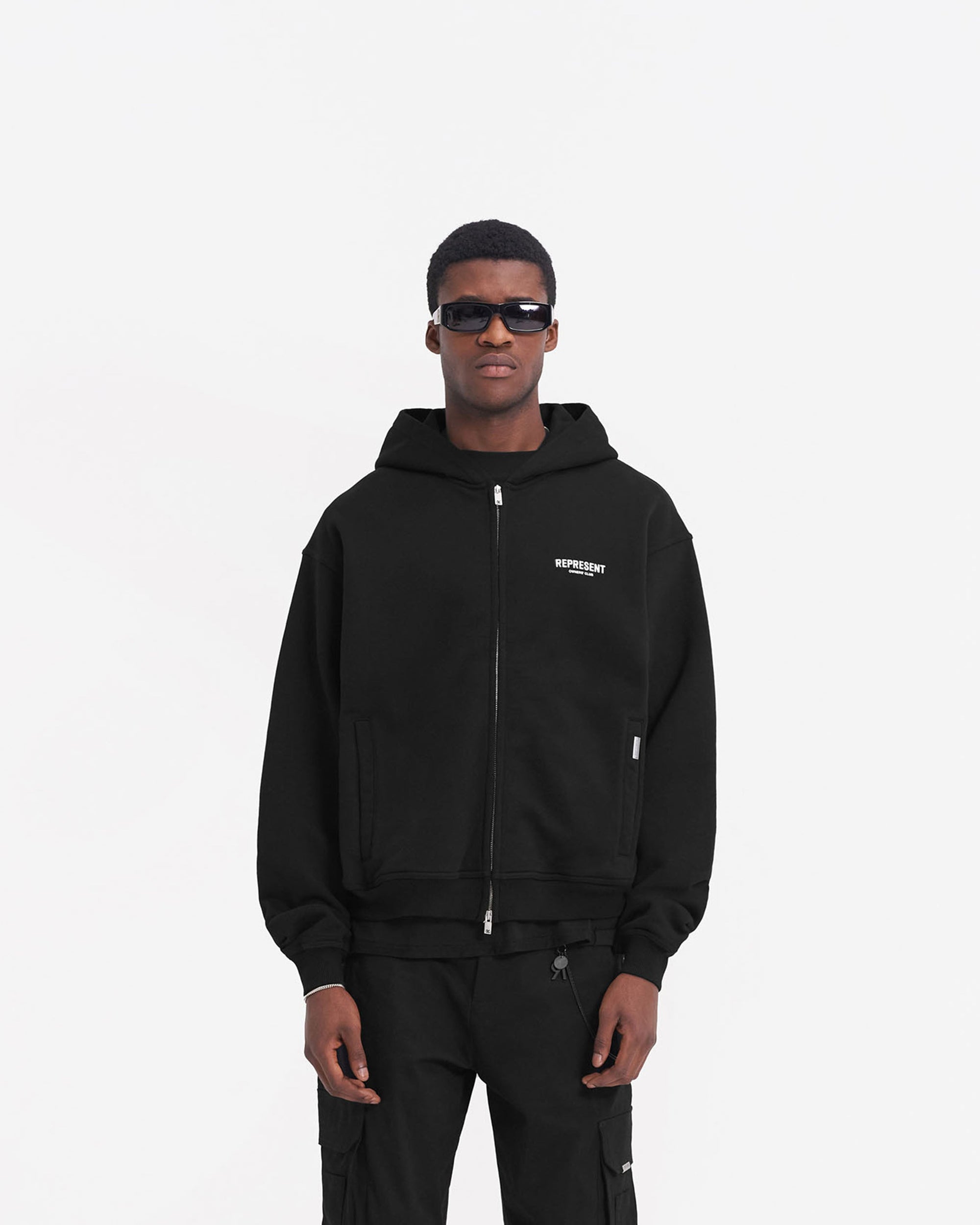 Man wearing a black zip hoodie on a plain white background | Represent Owners Club Zip Hoodie in Black | Oversized zip hoodie with ribbed trims | Owners Club Hoodies | Represent Clo