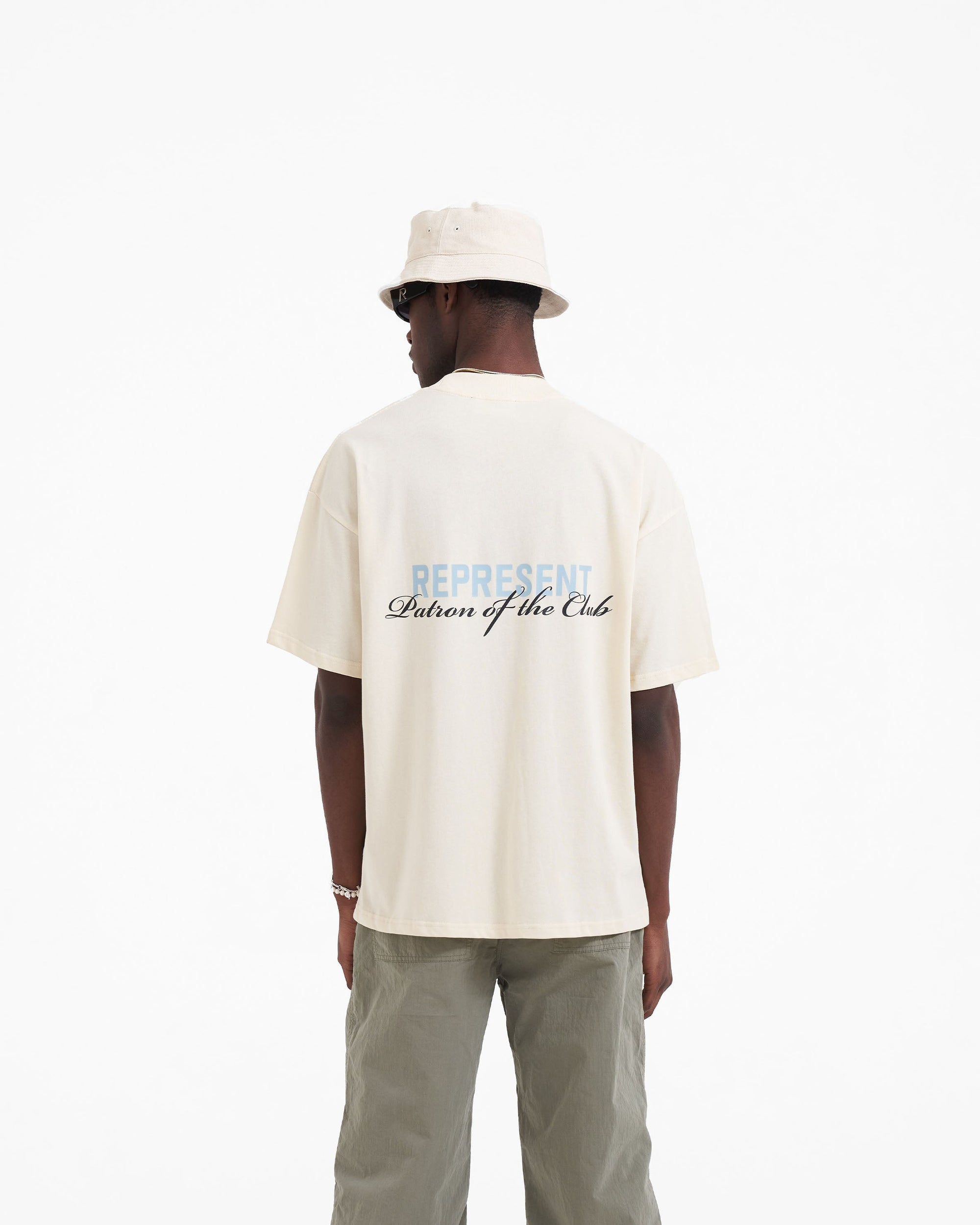 Man wearing a buttercream t-shirt on a plain white background | Patron Of The Club T-Shirt in Buttercream Powder Blue | Slightly oversized t-shirt with screen printed branding | Patrons T-Shirts | Represent Clo