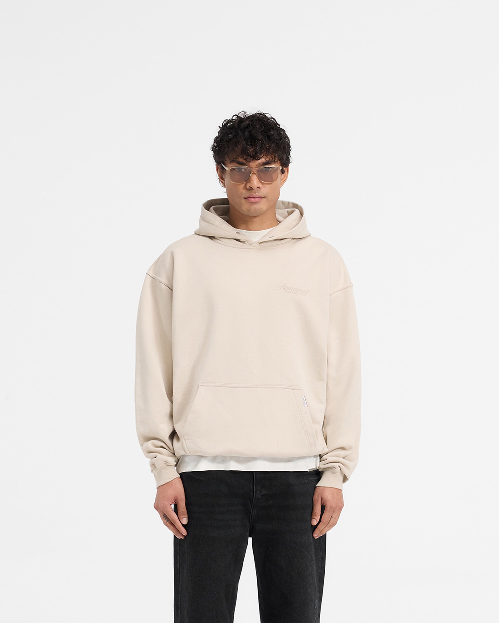 Man wearing an oversized grey hoodie on a plain white background | Represent X Ounass Owners Club Script Hoodie in Island Fossil | Heavy weight jersey cotton hoodie with screen print | Exclusives Hoodies | Represent Clo