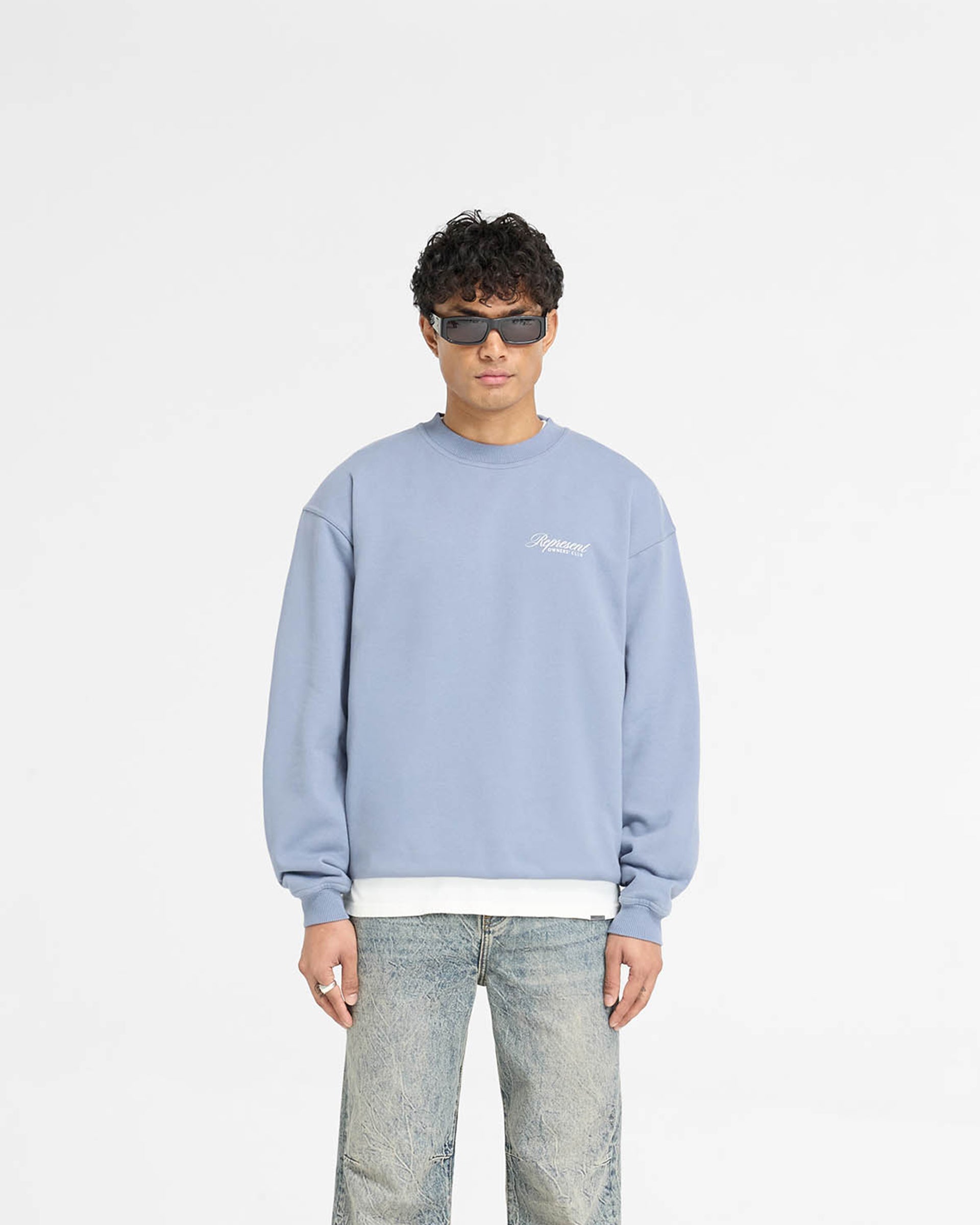 Man wearing a dusty blue sweater on a plain white background | Represent Owners Club Script Sweater in Dusty Blue | Oversized fit jersey sweater with ribbed trims | Owners Club Sweaters | Represent Clo