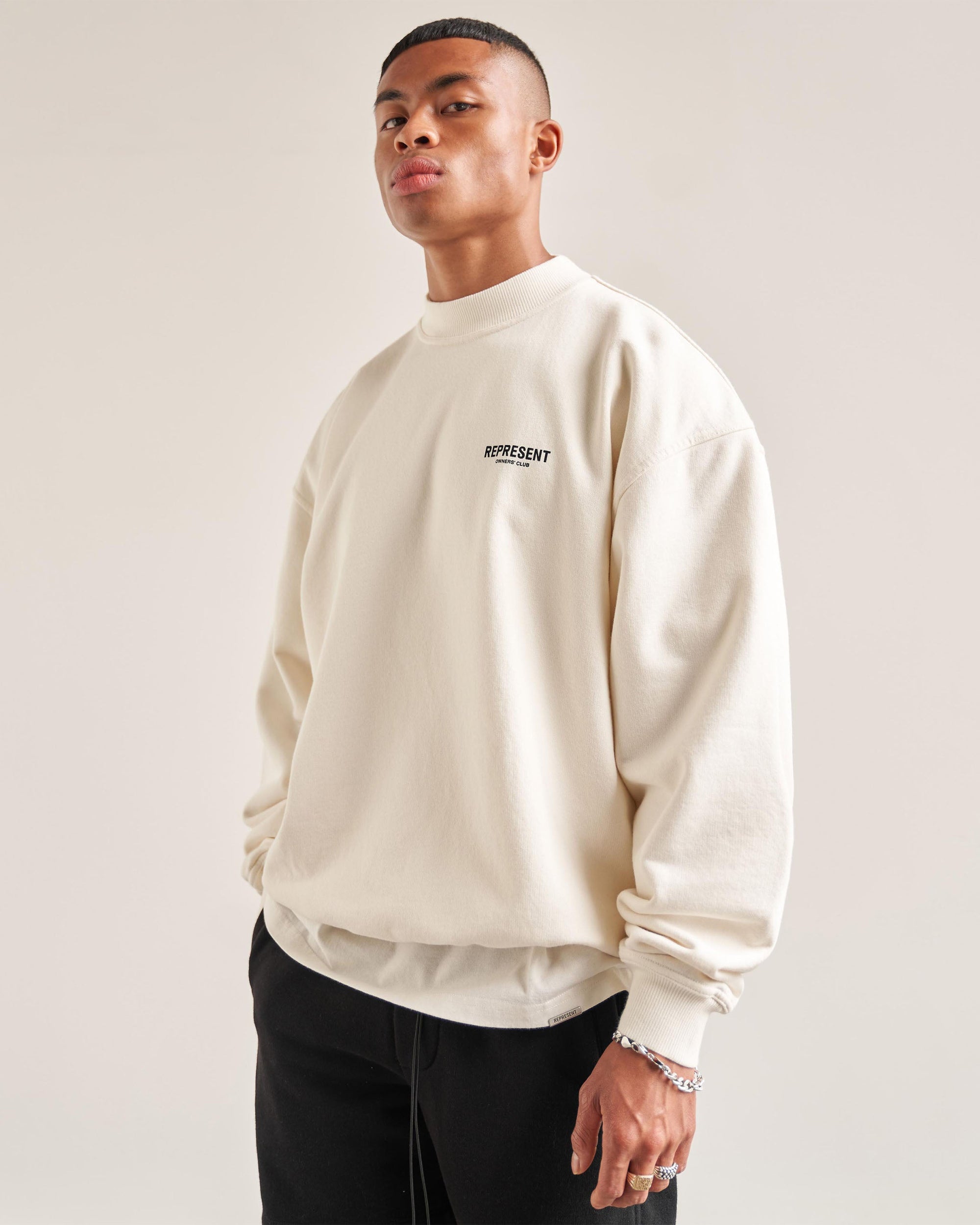 Man wearing a white sweater on a plain white background | Represent Owners Club Sweater in Flat White | Oversized fit crewneck sweater | Owners Club Sweaters | Represent Clo
