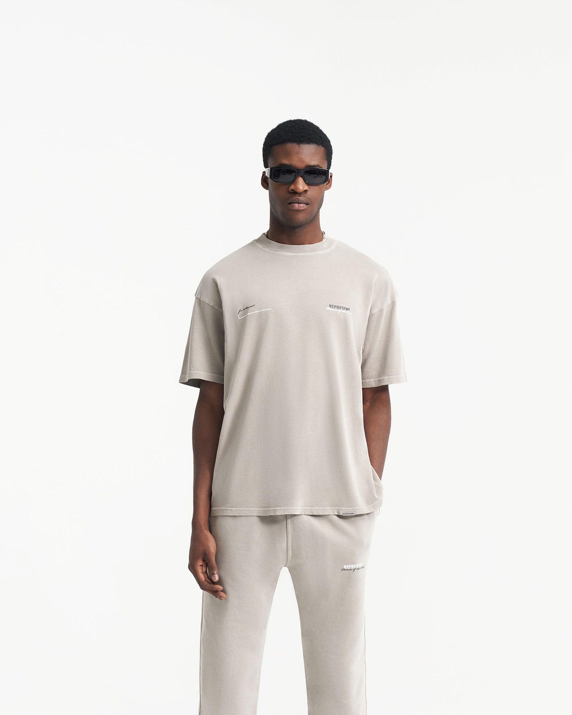 Man wearing an oversized grey t-shirt on a plain white background | Patron Of The Club T-Shirt in Mudstone | Short sleeve t-shirt with Patron of the Club branding | Patrons T-Shirts | Represent Clo