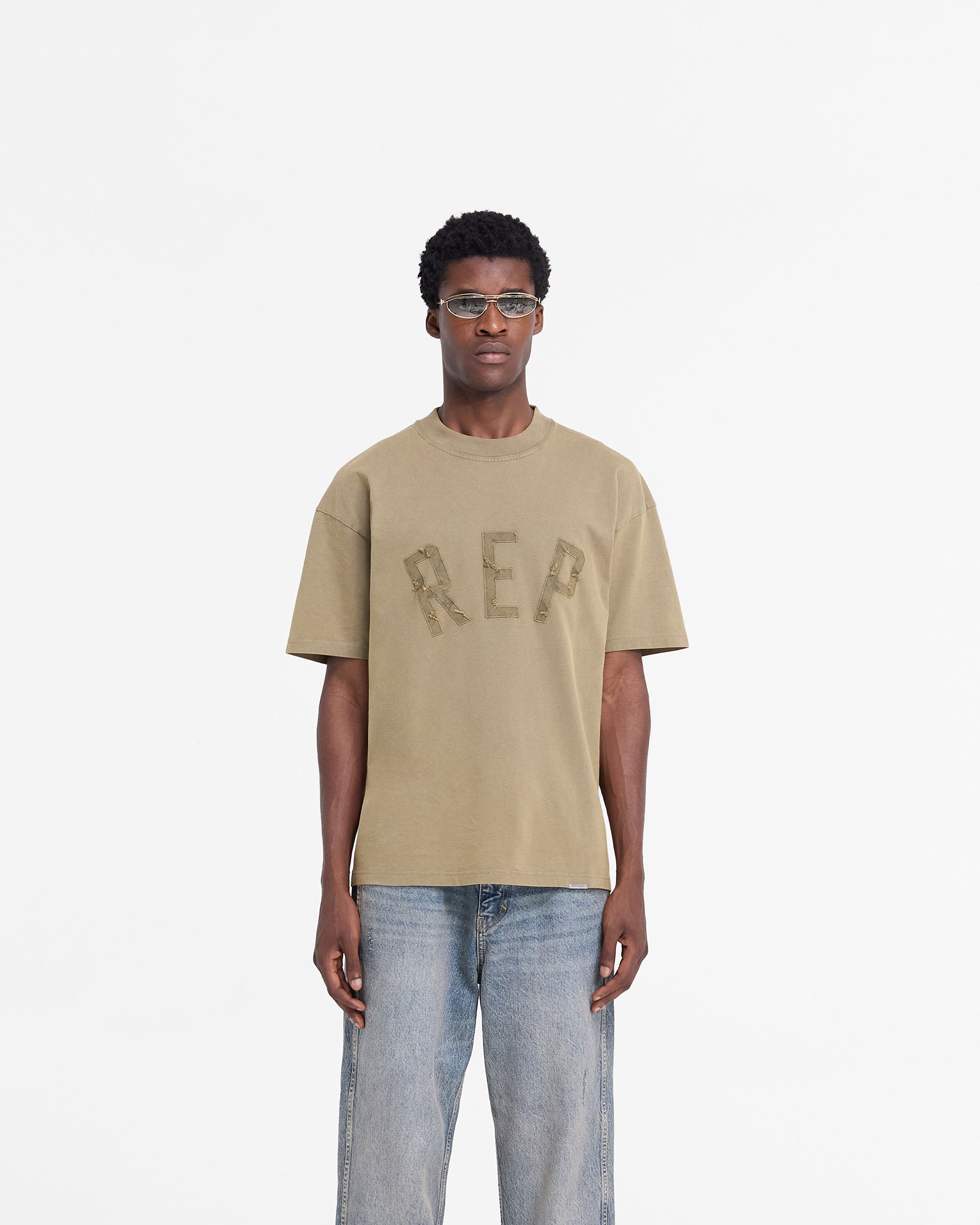 Man wearing a fawn t-shirt on a plain white background | Rep Applique T-Shirt in Fawn | Oversized t-shirt with distressed applique logo | FW24 T-Shirts | Represent Clo