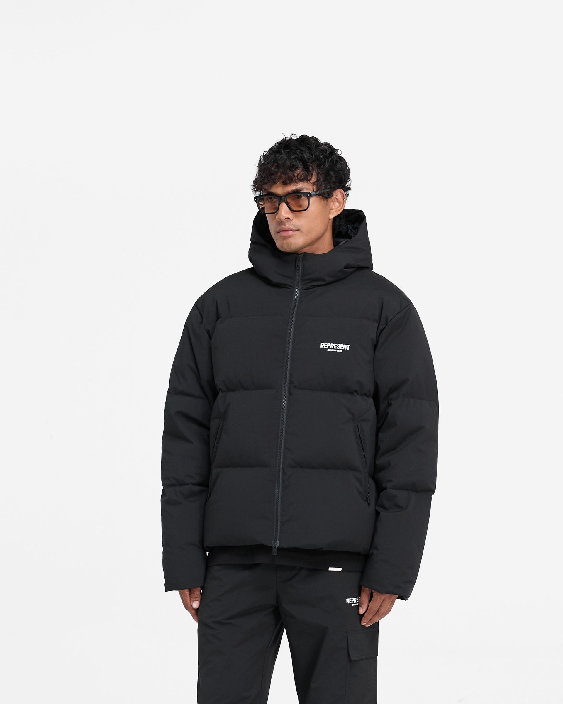 Man wearing a black hooded puffer jacket on a plain white background | Represent Owners Club Hooded Puffer Jacket in Black | Oversized duck down wadding puffer with zip pockets | Owners Club Outerwear | Represent Clo
